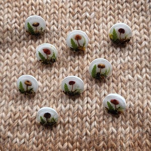 Könnte beinhalten: Neun weiße, runde Resin-Cabochons, jeweils mit einer Miniatur-Pilzszene. Die Pilze haben braune und rote Kappen, vor grünem Laub und Moos. Die Cabochons sind auf einem strukturierten, beigefarbenen Stoff ausgestellt.