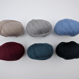 May include: Six skeins of yarn in various colors are arranged on a white surface. The colors include beige, gray, light blue, burgundy, teal, and black. The yarn is tightly wound into round balls, suitable for knitting or crochet projects.