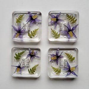 May include: Four square glass coasters with embedded purple flowers and green fern leaves. The coasters are clear with a grid pattern and are set against a white background. Each coaster features a unique floral arrangement.