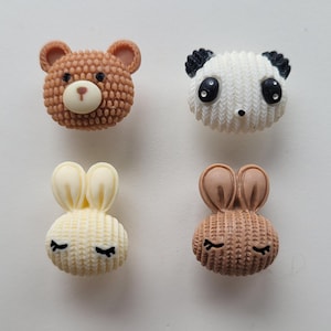 May include: Four different animal-shaped buttons with a knitted texture. The buttons are brown, white, and cream. The animals are a bear, a panda, and two rabbits.