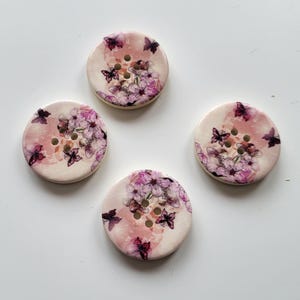 Op de afbeelding: Vier ronde knopen met een bloemen- en vlinderontwerp. De knopen hebben een crèmekleurige basis met een roze centrum. Het bloemmotief toont roze en paarse bloemen en vlinders.