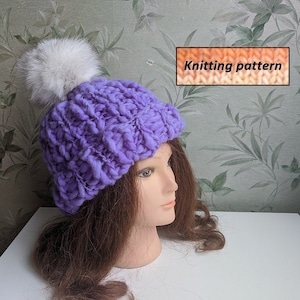 May include: A chunky knit lavender hat with a large white fur pom-pom. The hat is displayed on a mannequin head with brown hair. The image includes a text box that says "Knitting pattern".