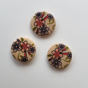 Op de afbeelding: Drie houten knopen met een bloemenpatroon. De knopen zijn beige met een roze, rood en zwart bloemenpatroon.