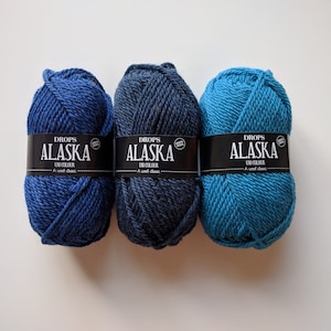 May include: Three skeins of Drops Alaska yarn in blue, dark blue, and teal colors. Each skein has a black label with white text that reads "Drops Alaska Un Colour A wool classic."