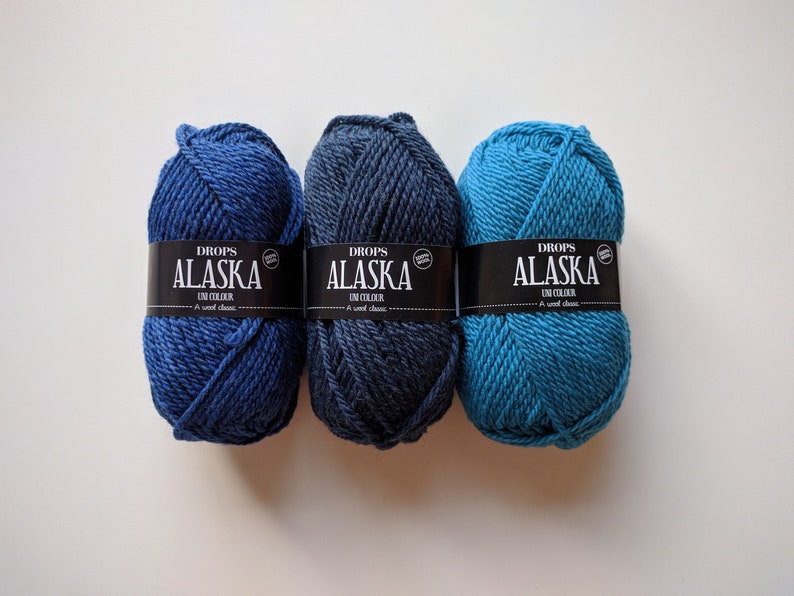 May include: Three skeins of Drops Alaska yarn in blue, dark blue, and teal colors. Each skein has a black label with white text that reads "Drops Alaska Un Colour A wool classic."