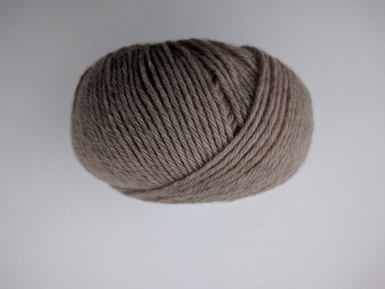 May include: A ball of taupe-colored yarn, tightly wound and textured. The yarn appears to be a neutral shade, suitable for various knitting or crochet projects. The image is a close-up, highlighting the yarn's texture and form.