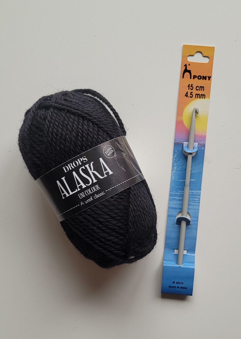 May include: A ball of black Drops Alaska wool yarn with a label that reads "Drops Alaska Uni Colour A wool classic 100% wool" and a gray crochet hook with a blue and orange label that reads "Pony 15 cm 4.5 mm #43410 Made in India"
