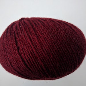 May include: A ball of burgundy yarn, tightly wound, against a white background. The yarn appears soft and textured, ideal for knitting or crocheting projects. The deep red color suggests warmth and richness.