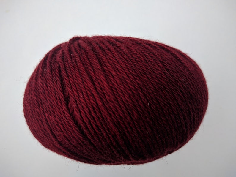 May include: A ball of burgundy yarn, tightly wound, against a white background. The yarn appears soft and textured, ideal for knitting or crocheting projects. The deep red color suggests warmth and richness.
