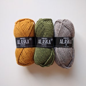May include: Three skeins of yarn in different colors: mustard yellow, forest green, and light gray. Each skein has a black label that reads "DROPS ALASKA" with the words "UNI COLOUR" or "MIX" underneath.