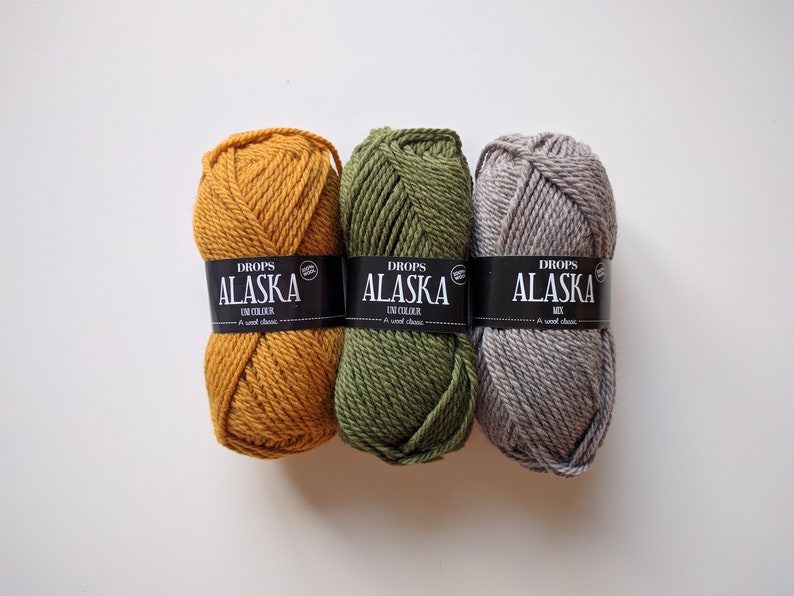 May include: Three skeins of yarn in different colors: mustard yellow, forest green, and light gray. Each skein has a black label that reads "DROPS ALASKA" with the words "UNI COLOUR" or "MIX" underneath.