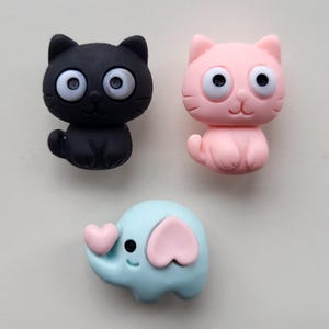 Op de afbeelding: Drie schattige knopen in de vorm van dieren: een zwarte kat, een roze kat en een lichtblauwe olifant met roze oren en een roze hart.