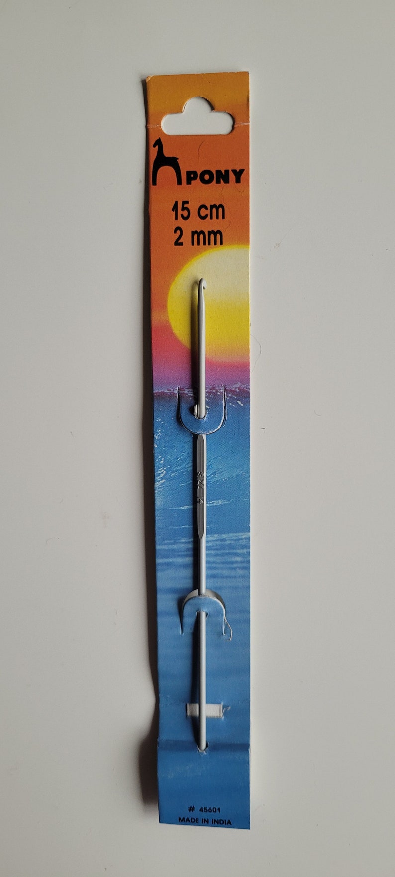 May include: A white Pony brand crochet hook with a 15 cm (6 inch) length and a 2 mm size. The hook is packaged on a blue and orange card with the brand name and size printed on it. The card also has a sunset scene with a yellow sun and blue water.
