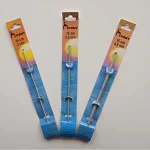May include: Three silver crochet hooks on their packaging. The hooks are labeled with the brand name "PONY" and the size in centimeters and millimeters. The packaging has a blue and orange sunset design.
