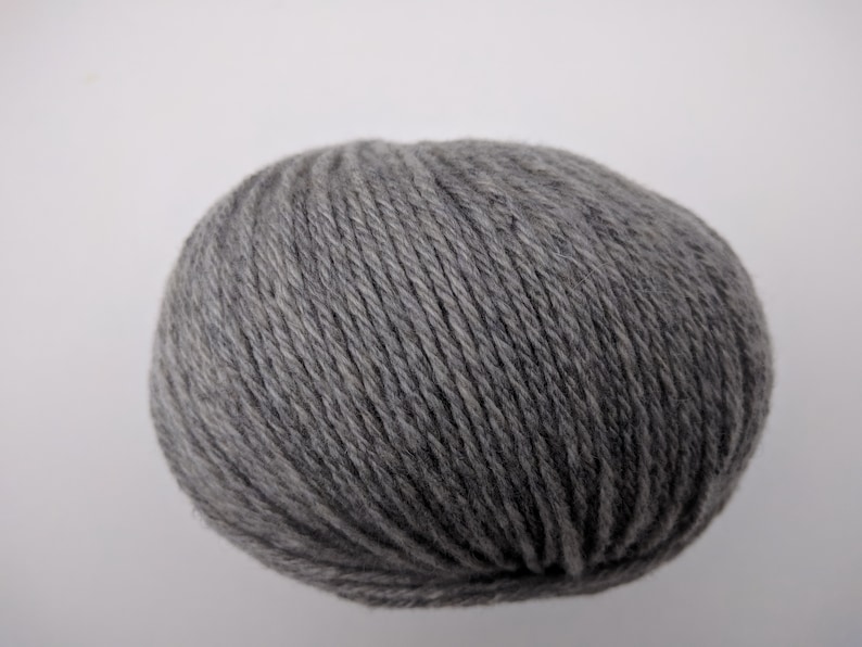May include: A ball of gray yarn, tightly wound and textured, sits against a plain white background. The yarn appears to be a medium weight, suitable for various knitting or crochet projects. The color is a neutral gray.