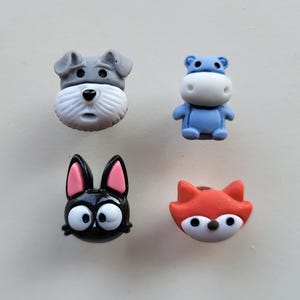 以下が含まれることがあります： 動物の顔が描かれたカラフルなプラスチックボタン4つ。ボタンはグレー、ブルー、ブラック、オレンジ。動物はシュナウザー、カバ、猫、キツネです。