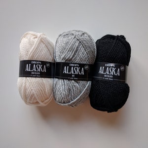May include: Three skeins of Drops Alaska yarn in white, gray, and black. Each skein has a black label with the text "Drops Alaska Uni Colour A soft classic".