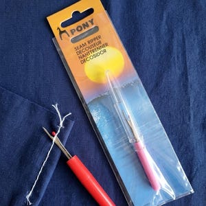 Könnte beinhalten: Ein roter und silberner Nahttrenner neben einem verpackten rosa Nahttrenner. Die Verpackung trägt den Markennamen "PONY" und die Worte "SEAM RIPPER DECOUSEUR NAHTTRENNER DECOSIDOR". Die Nahttrenner liegen auf einem blauen Stoff.