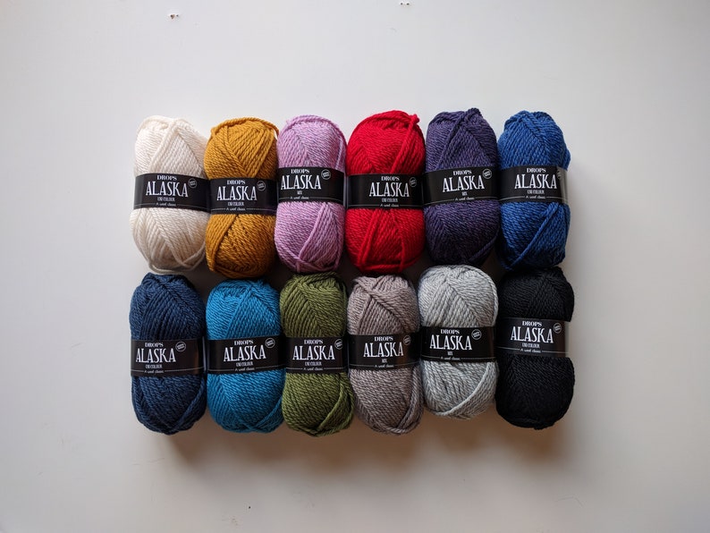 May include: Twelve skeins of yarn in various colors, including white, yellow, pink, red, blue, green, and gray. Each skein has a black label with the text "DROPS ALASKA" printed on it.