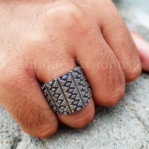 May include: A silver ring with a geometric pattern. The ring is wide and has a textured surface.