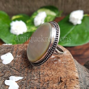 May include: A silver ring with a large oval-shaped mother-of-pearl stone set in a beaded bezel. The ring is on a wooden surface.