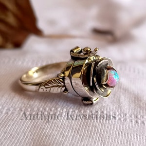 Könnte beinhalten: Ein silberner Ring mit einem rosafarbenen Fach, das sich öffnet und einen kleinen Raum freigibt. Die Rose ist mit einem rosafarbenen Opal verziert.