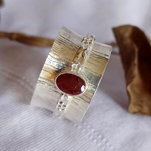 May include: A silver ring with a red gemstone set in the center. The ring is textured and has a decorative design around the band.