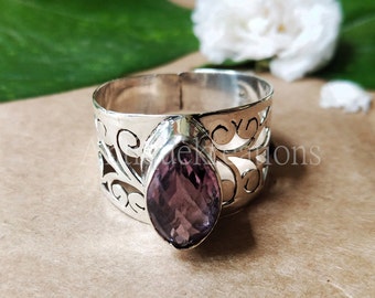 May include: A wide silver ring with an openwork design and a teardrop-shaped, purple gemstone. The ring's band features intricate cut-out patterns. The gemstone is a deep purple color, adding a touch of elegance.