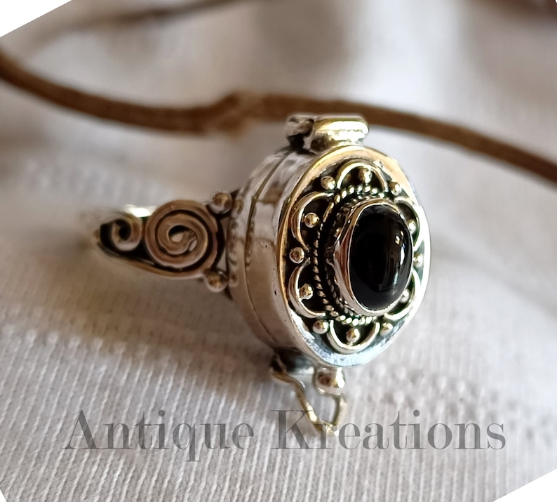 K&ouml;nnte beinhalten: Ein silberner Ring mit einem schwarzen Stein und einem Scharnierfach. Der Ring ist mit komplizierten Schn&ouml;rkeln und einem floralen Design verziert.