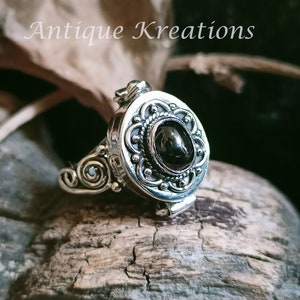 K&ouml;nnte beinhalten: Ein silberner Ring mit einem schwarzen ovalen Stein, der in einem filigranen Design gefasst ist. Der Ring ist &ouml;ffnungsf&auml;hig und hat ein kleines Scharnier an der Seite.