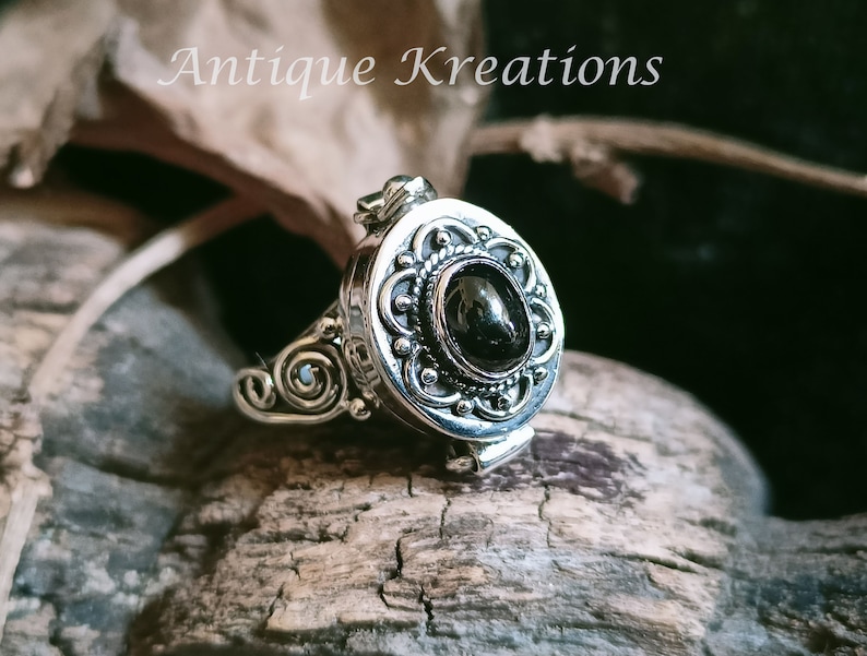 K&ouml;nnte beinhalten: Ein silberner Ring mit einem schwarzen ovalen Stein, der in einem filigranen Design gefasst ist. Der Ring ist &ouml;ffnungsf&auml;hig und hat ein kleines Scharnier an der Seite.