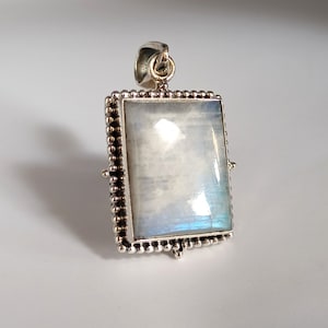 Pode incluir: Um pingente de prata com uma pedra de lua quadrada. O pingente tem uma borda de contas e uma argola de prata.