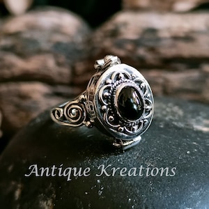 K&ouml;nnte beinhalten: Ein silberner Ring mit einem ovalen Anh&auml;nger, der mit einem schwarzen Edelstein besetzt ist. Der Anh&auml;nger hat komplizierte florale Designs und l&auml;sst sich &ouml;ffnen, um ein kleines Fach zu enth&uuml;llen.