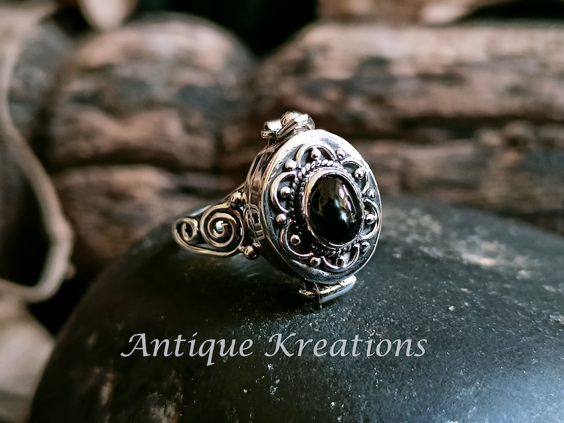 K&ouml;nnte beinhalten: Ein silberner Ring mit einem ovalen Anh&auml;nger, der mit einem schwarzen Edelstein besetzt ist. Der Anh&auml;nger hat komplizierte florale Designs und l&auml;sst sich &ouml;ffnen, um ein kleines Fach zu enth&uuml;llen.
