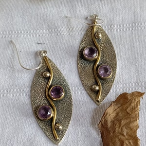 Puede incluir: Un par de pendientes de plata y oro con un diseño de hoja y tres piedras preciosas púrpuras. Los pendientes están sobre un fondo blanco.