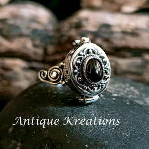 K&ouml;nnte beinhalten: Ein silberner Ring mit einem ovalen Anh&auml;nger, der mit einem verzierten Design und einem schwarzen Edelstein verziert ist. Der Anh&auml;nger l&auml;sst sich &ouml;ffnen und zeigt ein kleines Fach.