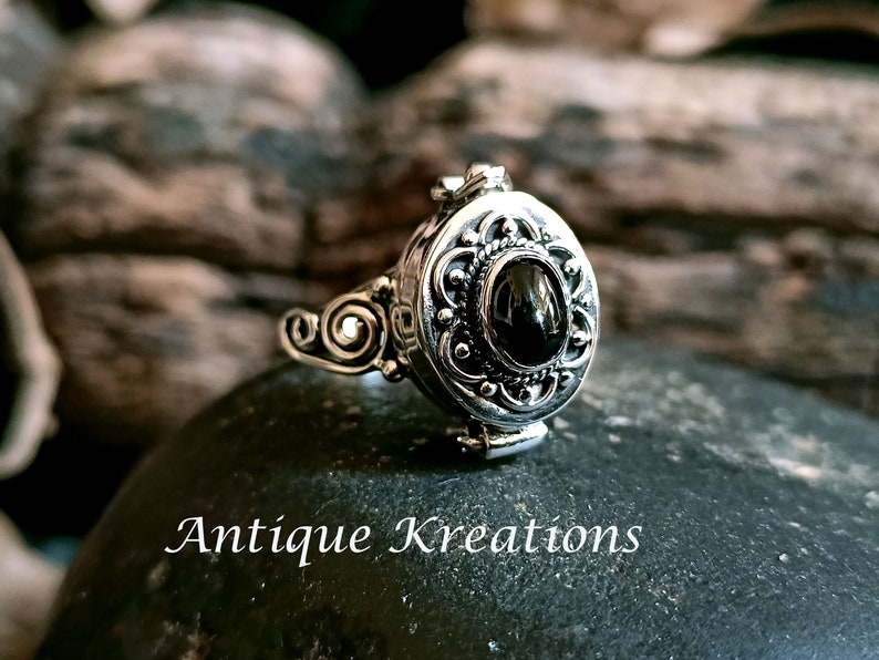 K&ouml;nnte beinhalten: Ein silberner Ring mit einem ovalen Anh&auml;nger, der mit einem verzierten Design und einem schwarzen Edelstein verziert ist. Der Anh&auml;nger l&auml;sst sich &ouml;ffnen und zeigt ein kleines Fach.