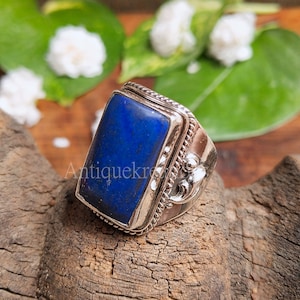以下が含まれることがあります： 銀のリングに、大きな長方形の濃い青色のラピスラズリが、細工の施された銀のベゼルにセットされています。リングの側面には、装飾的な銀のデザインが施されています。