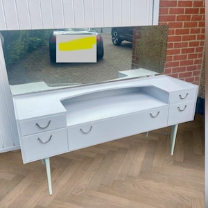 May include: A white vanity table featuring a large rectangular mirror, a central open storage shelf, and four drawers with silver-toned curved handles. The piece is supported by four tapered legs and has a smooth, clean finish throughout the structure.
