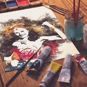May include: A watercolor painting of Wonder Woman on a white sheet of paper. The painting is surrounded by paint tubes, brushes, and a jar of water. The paint tubes are labeled with the brand name "Winsor & Newton" and the color name.