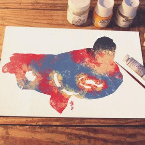 May include: A watercolor painting of Superman, with red, blue, and yellow paint splatters. The painting is on a white canvas, and there are paint tubes and brushes on a wooden table.
