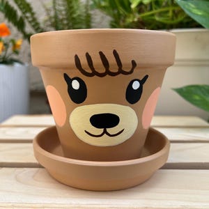 May include: A brown terracotta flower pot with a matching saucer. The pot is hand-painted with a cartoon bear face, featuring black eyes, a black nose, and peach-colored cheeks. The pot is designed for planting flowers or herbs.