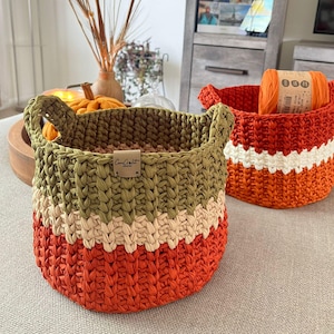 May include: Two handmade crochet baskets. One is orange, cream, and olive green, with handles and a label that reads "CanCraft". The other is orange with a cream stripe. A ball of orange yarn sits inside.