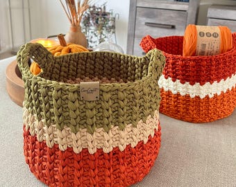 May include: Two handmade crochet baskets. One is orange, cream, and olive green, with handles and a label that reads "CanCraft". The other is orange with a cream stripe. A ball of orange yarn sits inside.