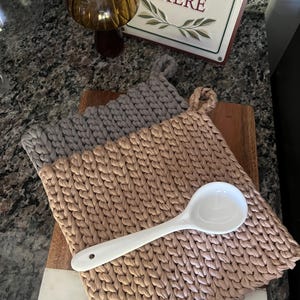 Może przedstawiać: Dwa ręcznie robione, dziergane podkładki pod garnki w kolorze beżowym i szarym, z białą ceramiczną chochlą na wierzchu. Podkładki pod garnki są kwadratowe z pętelką do zawieszania. Tło obejmuje drewnianą deskę do krojenia i znak z napisem "Gather Here".