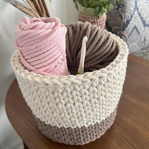 May include: A crocheted basket with a white and brown striped design, filled with pink and brown yarn and a crochet hook.
