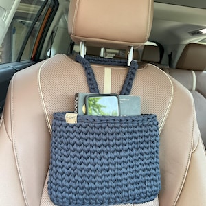 May include: A dark blue, crocheted car organiser bag suspended from a car headrest. The bag contains a phone and a book. The bag has a small wooden tag with text. The car seat is brown.