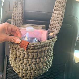 May include: A green crocheted hanging basket with a brown leather tag, attached to the headrest of a car seat. The basket contains a clear water bottle and a pink and white book.