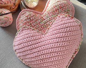 May include: Two pink crocheted heart-shaped cushions. The top cushion has a floral pattern and the bottom cushion is a solid pink.
