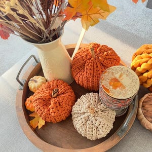 May include: A wooden tray displays orange and cream-coloured crocheted pumpkins, a small candle, and a cream-coloured jug with autumn foliage. The pumpkins vary in size and are arranged with autumn leaves.